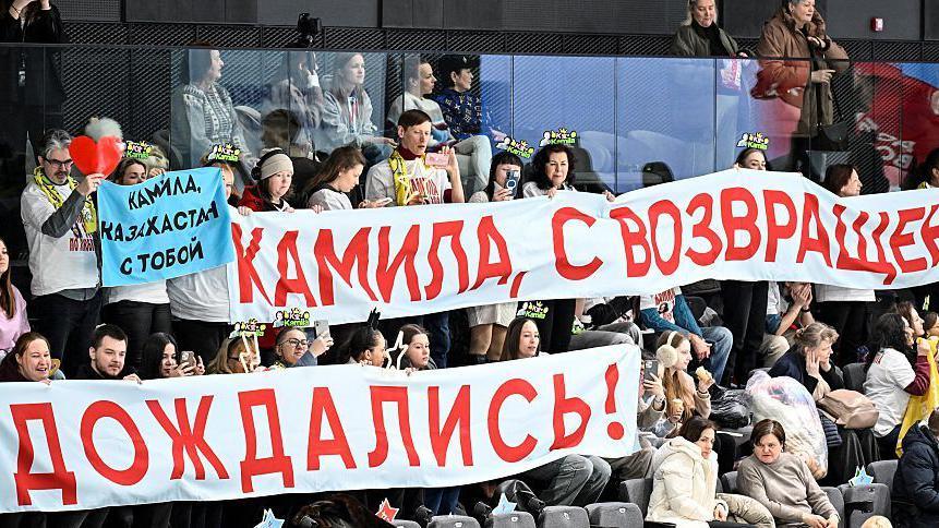 People in the crowd hold up two large white banners which have Russian writing on them. A man and woman are also holding a smaller blue banner in support of the skater.