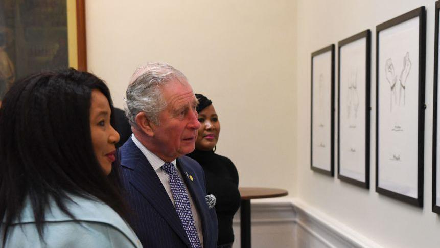 Makaziwe Mandela, who has long black hair and wears a blue jacket, looks at her father's artworks while standing next to Prince Charles during a visit to St George's Hall. There are three artworks up including Imprisonment with the hands tied.