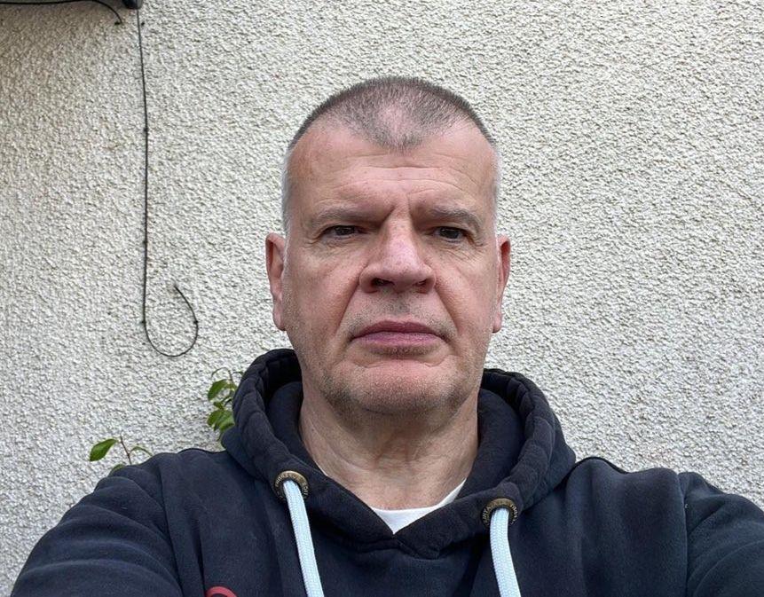 John Meiklejohn, a middle aged man dressed in a dark coloured hoodie, looks at the camera with a rendered wall of his home in the background.