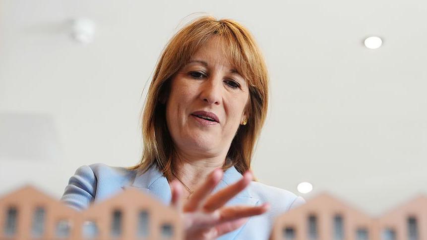 Chancellor Rachel Reeves, wearing a powder blue suit, standing with her hand over some model houses which are just seen in front of her