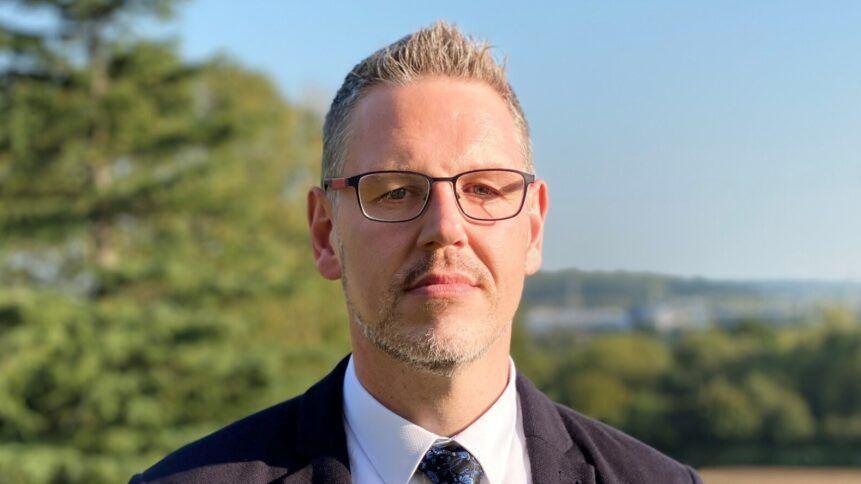 The West Mercia Police and Crime Commissioner John Campion, a man with short grey hair, a short grey beard and glasses. He is wearing a suit and tie and is standing outside.