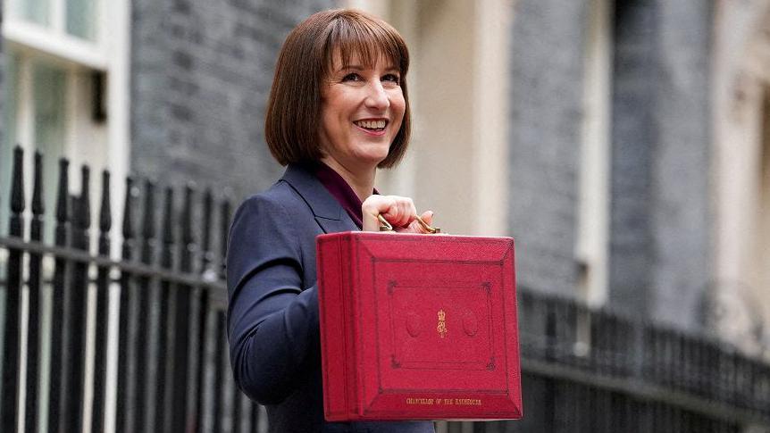 Chancellor Rachel Reeves standing outside Downing Street holding up a red briefcase that says "Chancellor of the Exchequer" and the Royal cypher. In gold writing. Rachel Reeves is smiling, with short brown hair and wearing a navy suit.