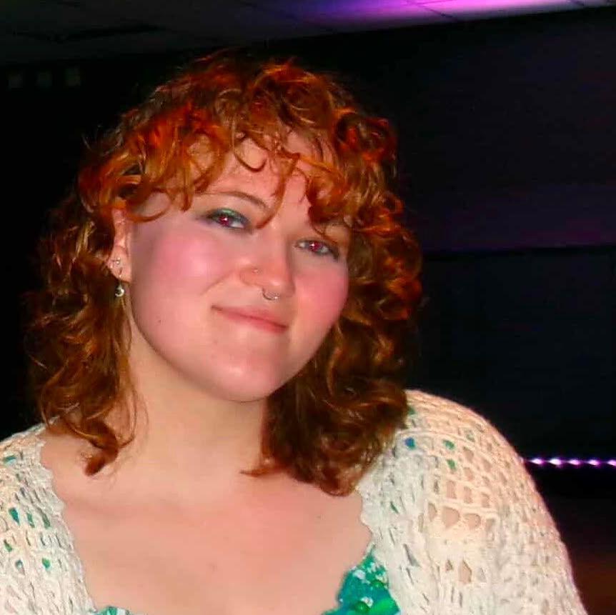 A portrait photograph taken indoors in a dark entertainment venue. One person is positioned in the centre of the frame, facing the camera from slightly above waist height. The person has shoulder-length curly red hair and is wearing a white open-knit cardigan over a green-and-white top decorated with a leafy botanical pattern. Small facial piercings are visible on the nose and ears. The lighting is low, with purple and pink light cast across the ceiling and faintly visible in the background. Most of the background is dark, making the person stand out against the surroundings. 