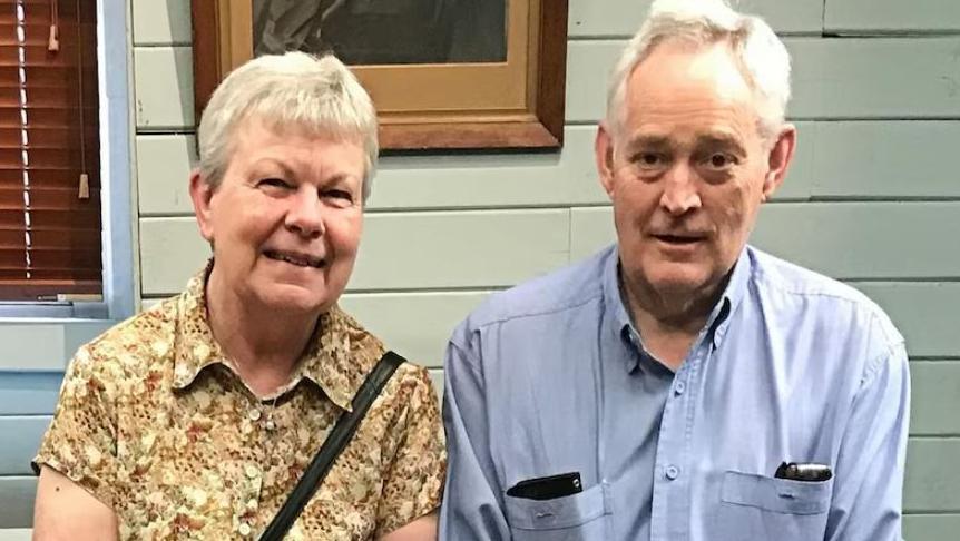 Heather and Ian Wilkinson smile at the camera, while sitting in a room. Heather is wearing a yellow patterned shift while Ian is wearing a blue shirt with his phone and glasses each tucked into his shirt pockets.