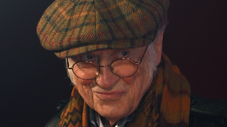 Noddy Holder against a dark background with a brown cloth cap and scarf