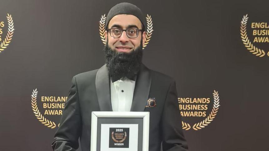 A man with glasses, a long black beard and wearing a tuxedo, smiles while he holds a framed certificate. He is standing in front of a black wall which says "England's Business Awards" in gold lettering.