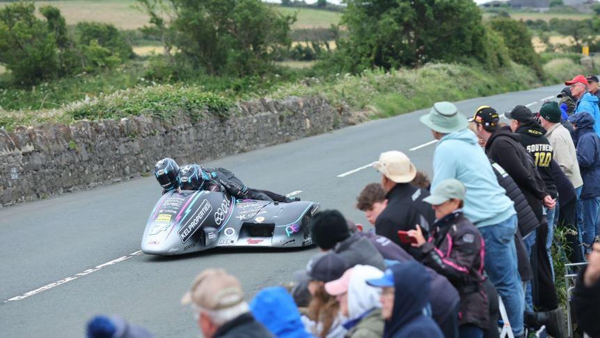 A sidecar outfit on the track with a stone wall on the left, and spectators leaning over another on on the right.