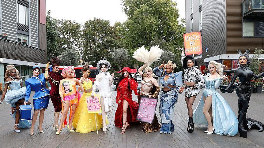 12 drag queens stood in a line posing, they are all wearing bright bold costumes each one is different.