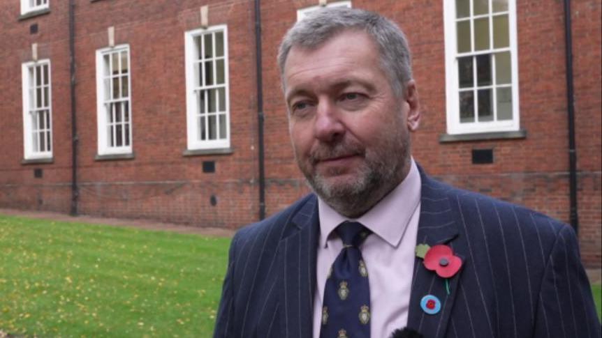 A man with short grey hair and a black blazer with a navy tie and a remembrance poppy is stood near grass outside a brick building. He is looking away from the camera to the side.