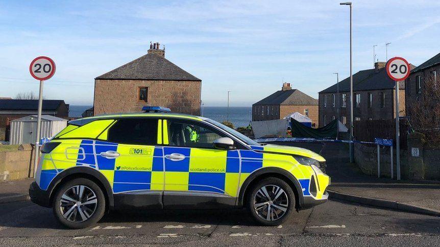 Police car with yellow and blue markings at scene of incident, with police tape in background, and two 20mph speed limit signs, and blue sky.