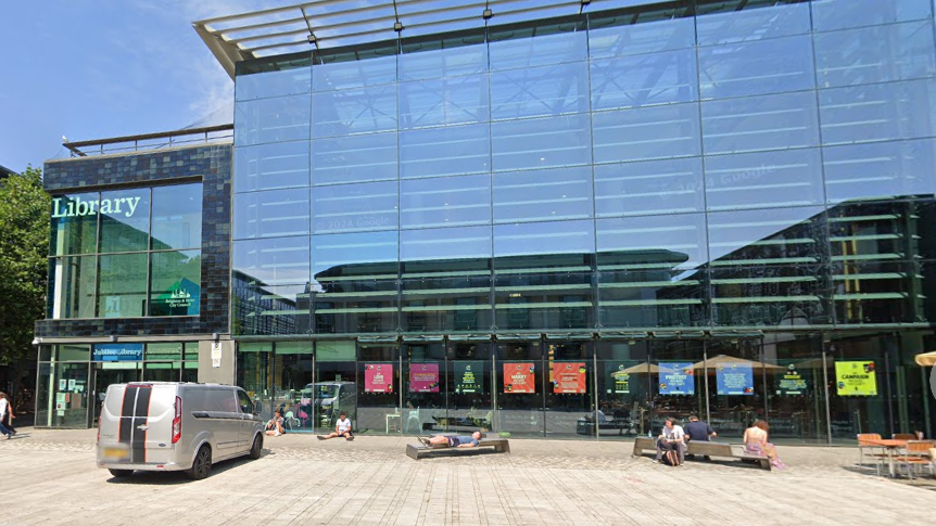 A large building with people sitting outside and a grey van.