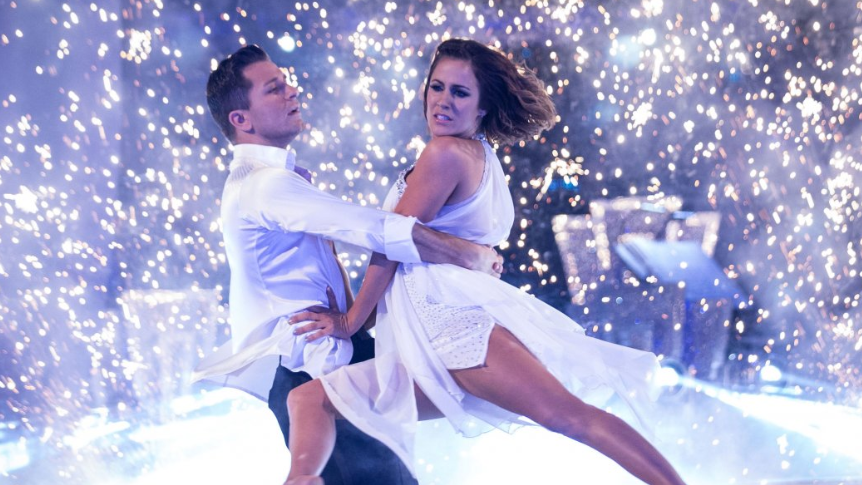 Pasha Kovalev, in a white shirt and dark trousers and Caroline Flack, in a white floaty dress, in mid-dance, with Caroline lifted by Pasha as sparkling white lights form a backdrop