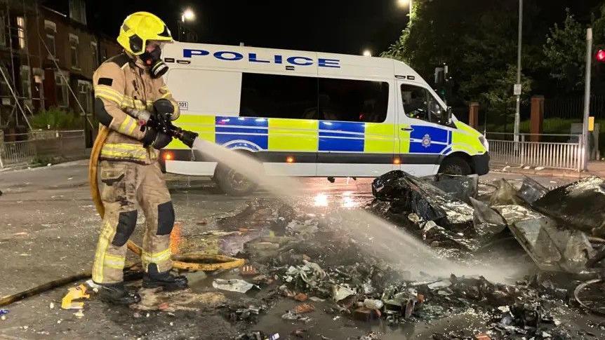 A firefighter extinguishes flames amid a pile of rubble on a street. A marked police van stands in the background. It is night time. 