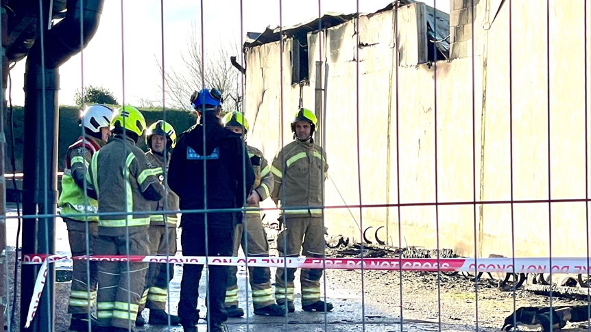 Firefighters in uniform and helmets standing together, in discussion, behind a fire cordon and metal fence, beside a fire-damage farm building