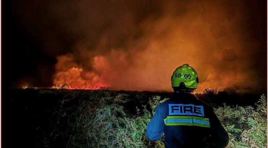 The image shows a firefighter standing in front of a massive wildfire at night on Dartmoor. They're wearing full protective gear, including a helmet marked "5000" and a jacket with "FIRE" on the back. The flames are towering and fierce, lighting up the sky with an intense orange glow. Smoke billows upward, and the ground is covered in vegetation, suggesting the fire is sweeping through grassland.