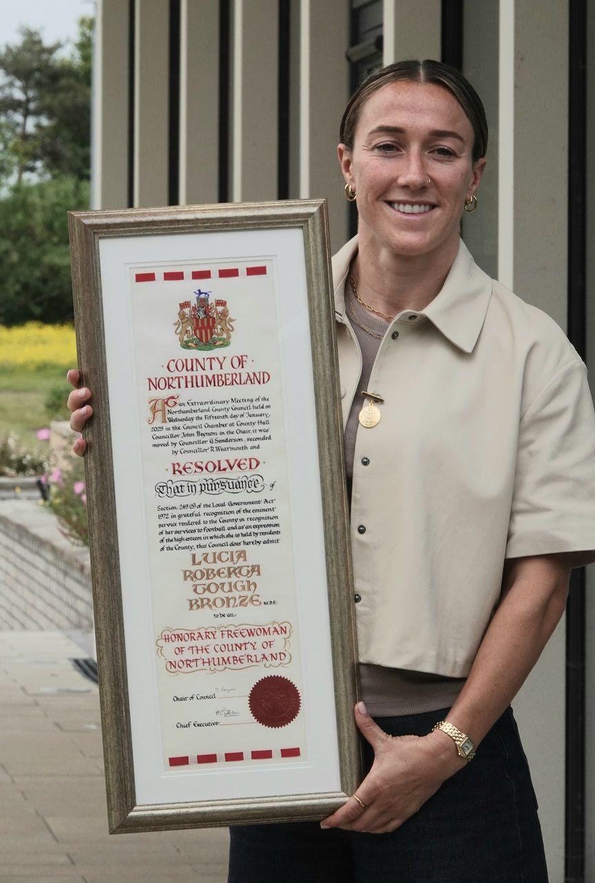 Lucy Bronze holds a long certificate which is the official Freedom of Northumberland. Her name is on it and it has very traditional writing, with the Northumberland county coat of arms. She is smiling and wears a smart cream-coloured jacket.