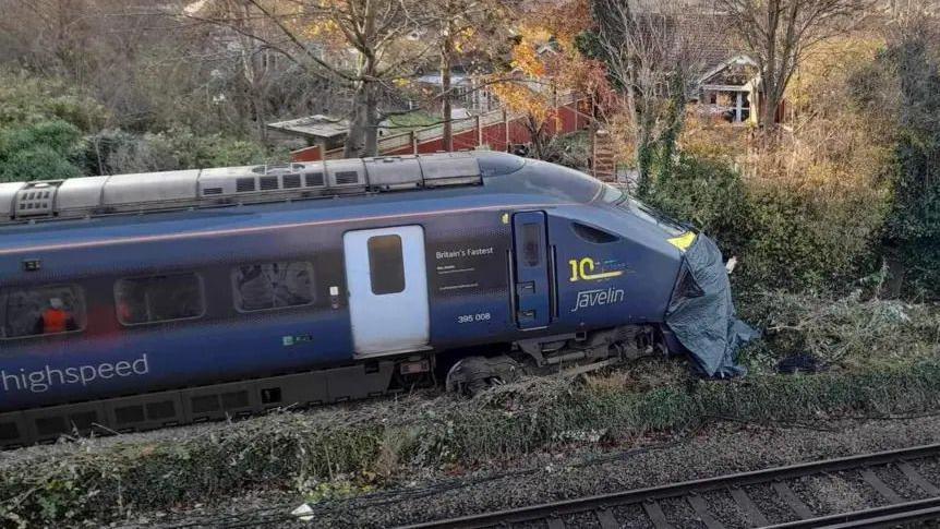 A blue train with the words high speed written on the side in grey. The train has left the track and is in green undergrowth. Trees are in the background