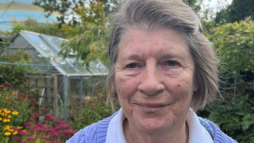 Smiling Pat Longton standing in her garden surrounded by flowers and trees with a greenhouse in the background