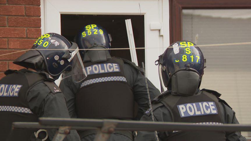 Three police officers enter a house. They are wearing helmets and other protective clothing.