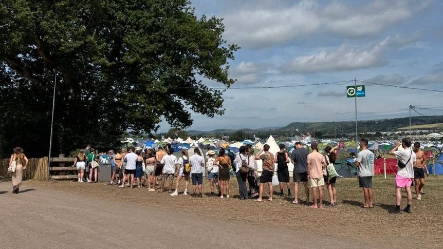 A queue of people waiting for their turn to fill up their water supply. There are tents in the background.