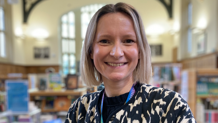 The picture shows Sarah Herlinger smiling at the camera. She is standing in the school's library