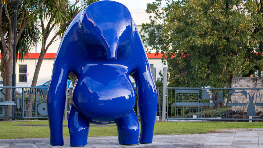 A large blue animal-like sculpture with long arms and short legs, an extended belly and a faceless head with a snout-like nose