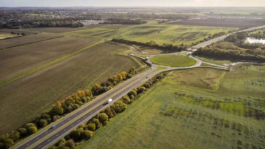 Birds eye view of the M181 junction in North Lincolnshire. There are two lanes with trees either side leading up to a roundabout surrounded by fields.