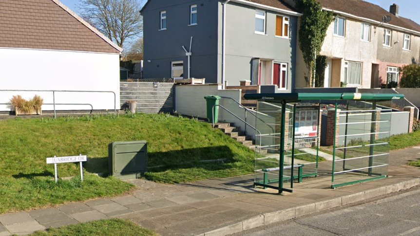 A pavement along a road which has a bus stop on it and a grassy raised verge with a sign that reads UXBRIDGE DR. There is a block of houses behind the bus stop.