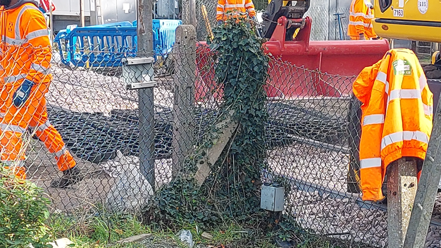 A metal fence in front of a railway crossing on which sections of black rubber have been placed by workers wearing orange high-vis clothing