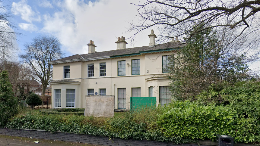 A large, whitewashed villa-style building is surrounded by trees and other greenery