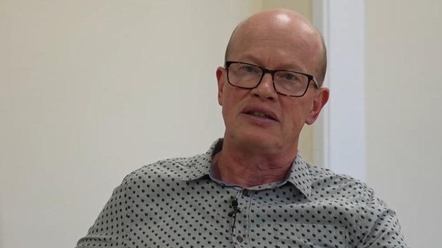 Paul de Kort, who is bald and wearing tortoise-shell glasses and a grey shirt with black dots. He is sat inside a cream room and has his mouth slightly open, as if he is talking.