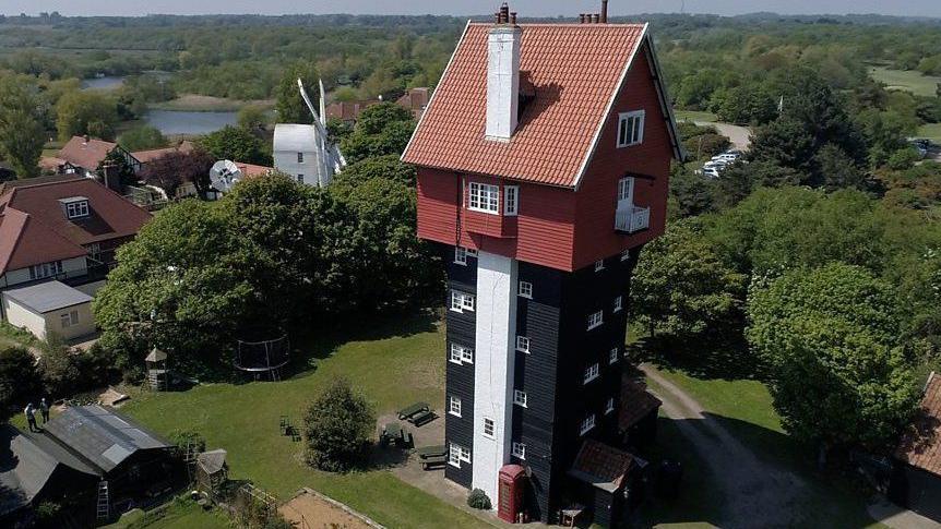 The House in the Clouds at Thorpeness. The former water tower has a black clad five storey tower base, above which is a house with red walls and roof. The windows of the building are all painted white. Behind it can be seen a white windmill, the roofs of houses in amongst trees and beyond that a lake on the left