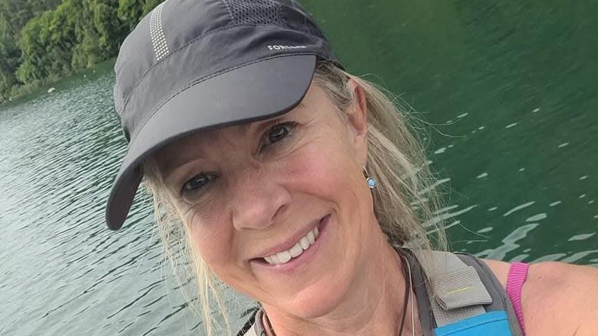 Kath Maguire of The Feel Good Factor club, with blonde hair pulled back behind her head, wearing a black cap and safety jacket, there is water in the background.