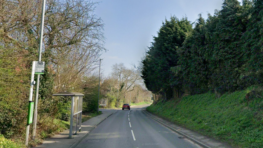 A Google Streetview image of New Mill Lane, which is a tarmac road with one lane in either direction. There is a bus stop with metal and glass shelter to the left and a grassy verge and trees to the right
