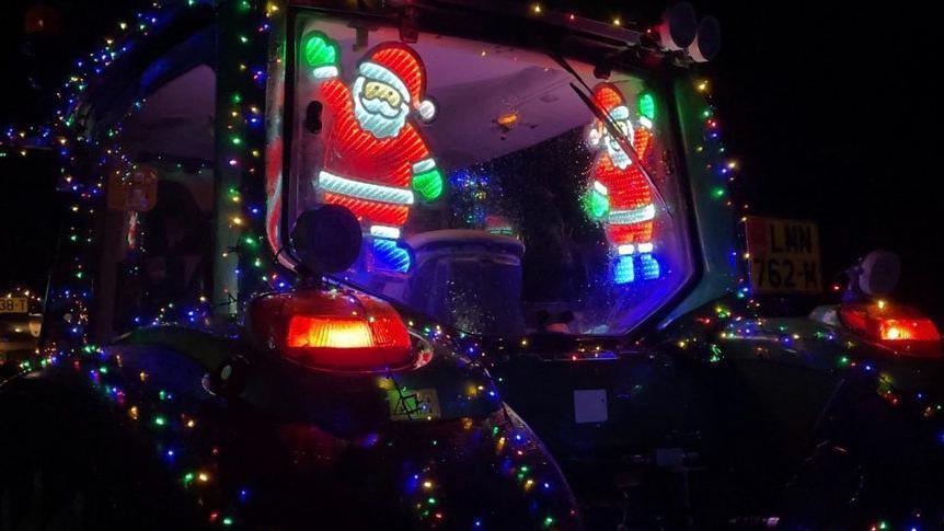 The rear window of a tractor's cabin with light-up Santa figurines. The exterior of the tractor is also decorated with illuminated multi-coloured Christmas lights decorating the tractor's exterior.