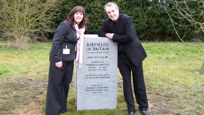 Kenneth Bannerman is leaning on the grey stone memorial which reads "Airfields of Britain Conservation Trust... in memory of Fambridge Airfield". He is wearing a black suit and black shoes. On the other side of the memorial is a woman wearing a black pinstripe suit, pink scarf and a white name badge. 