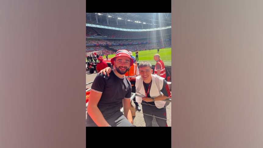One man is looking at the camera, wearing a red bucket hat and black t-shirt. Another man is bending towards him, with his arm around his shoulder. Both are posing as one sits, and the other stands in a football stadium. A football pitch can be seen behind them.