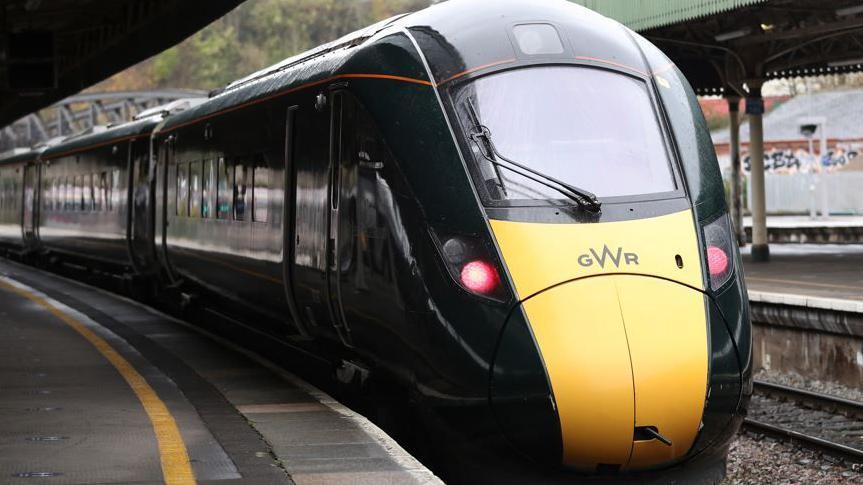 A green train with a yellow nose with the letters GWR in a train station.