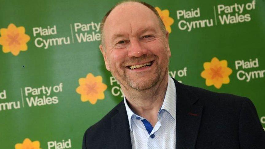 A smiling Cefin Campbell wearing a light shirt open at the neck and a dark jacket. Behind him is a large green hoarding panel with the words Plaid Cymru Party of Wales printed in white lettering alongside the party's yellow poppy emblem.