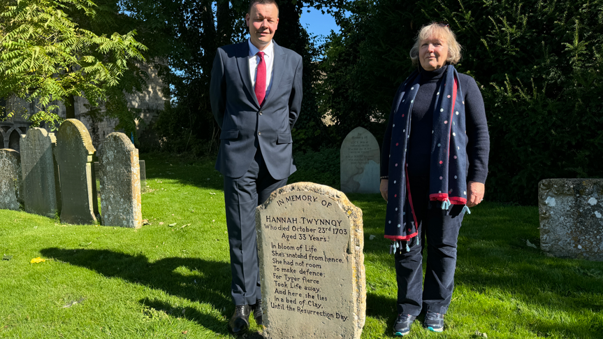 Chris Brooks and Cristina Staff standing smartly dressed in sunshine behind the headstone in the grounds of Malmesbury Abbey. 