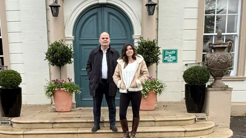 A man and women standing side-by-side on steps leading up to the door of a stone building. He is bald and is wearing a black coat, white shirt and black shoes. She has long brown hair and is wearing a white top, black jeans, brown boots and a light brown jacket.