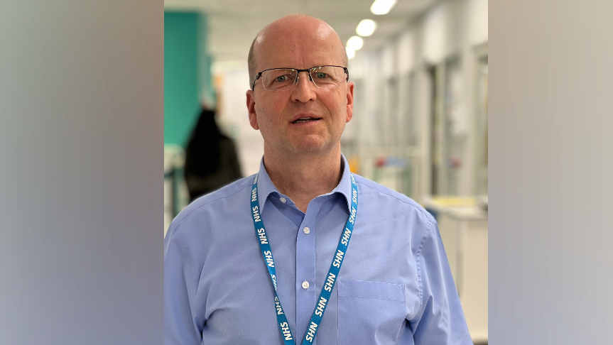 A bald man with glasses looks into the camera, standing in a hospital corridor, wearing a blue shirt and an NHS blue lanyard