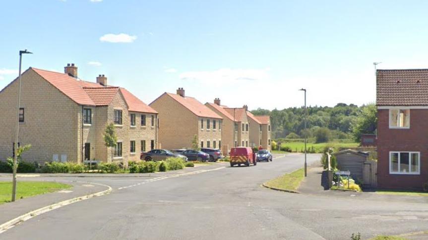 A smart-looking residential street runs down towards green fields and trees under a blue sky. Cars are parked in the drives of modern yellow-brick houses. A post van is parked in the street. 