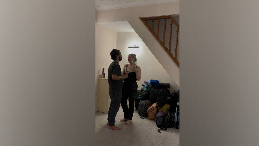 A man and woman, dressed in dark clothing, hold two wine glasses as they cheers each other. Both are standing in an empty living room, lit by a wall light and surrounded by bags and backpacks.