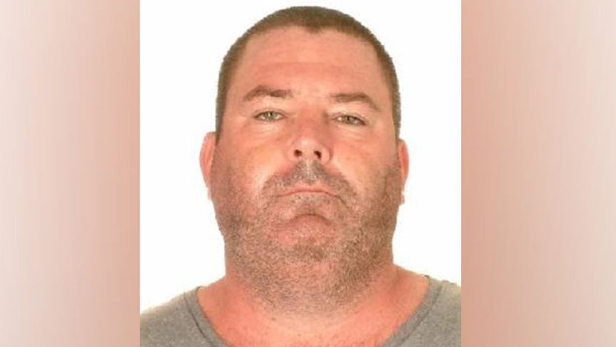 A police mugshot of a man wearing a grey t-shirt. He has very close-cropped short brown hair and stubble. He has a chubby face and neck of roughly the same width and appears to be sunburnt. 