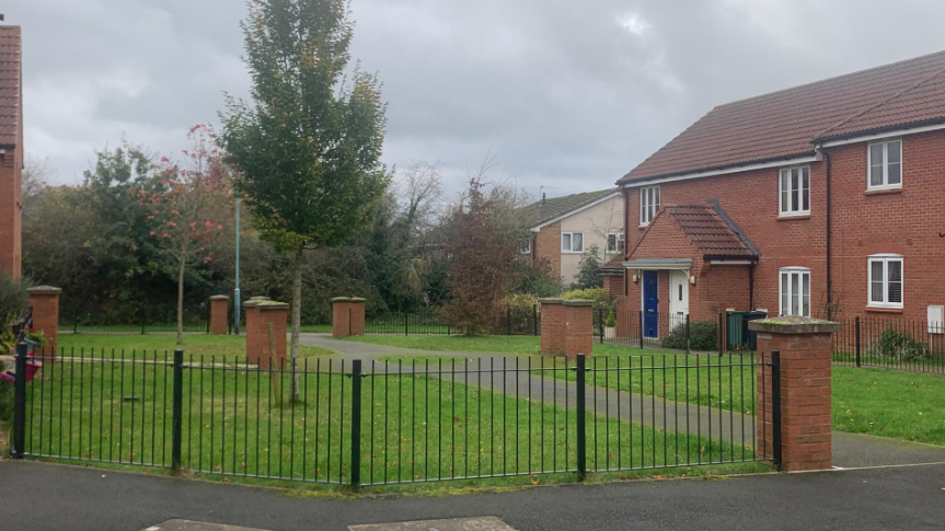 An image of an open area in front of a red brick house, with the area enclosed by a black fence