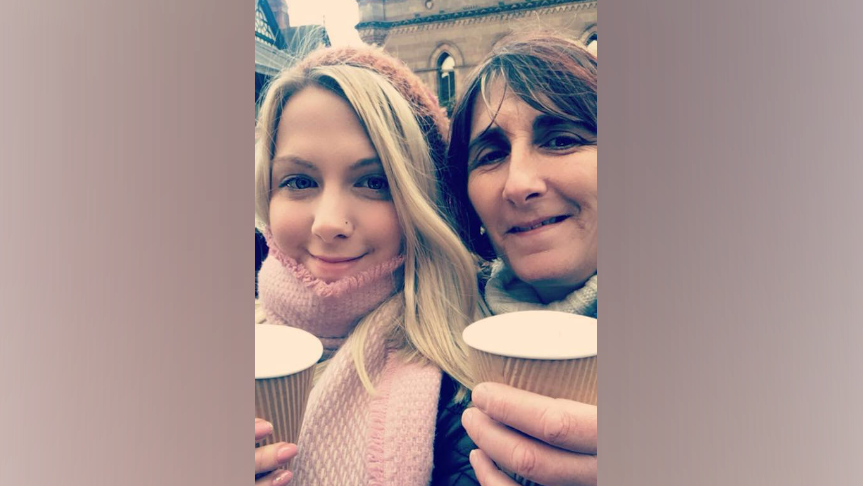 A young girl with blonde hair and a hat, and a pink scarf, holds a cup, next to an older woman with reddish brown hair, also holding a cup, both are smiling into the camera 