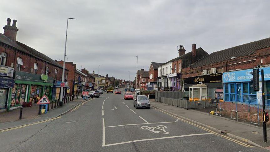 A road in Leeds with shops on either side and some cars. Traffic lights and a road sign can be seen.