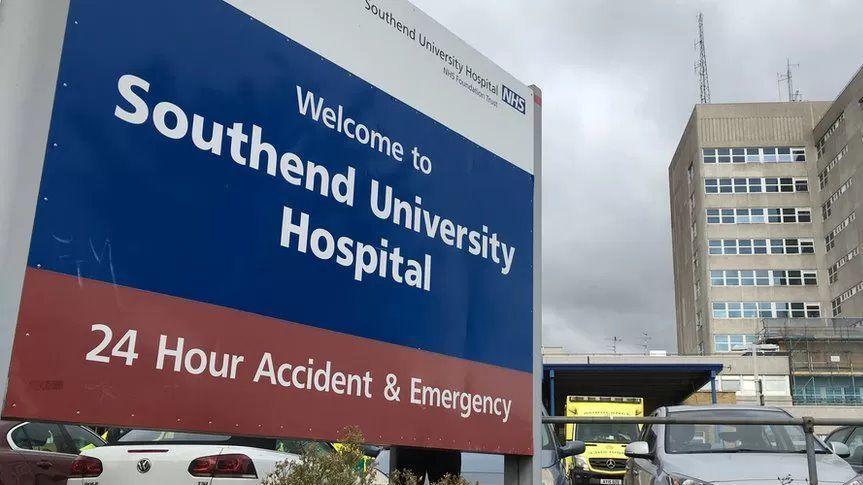 A sign at the entrance to Southend Hospital, showing where the accident and emergency unit is. In the background is a large grey building, as well as cars and an ambulance. The picture has been taken on a grey, cloudy day.