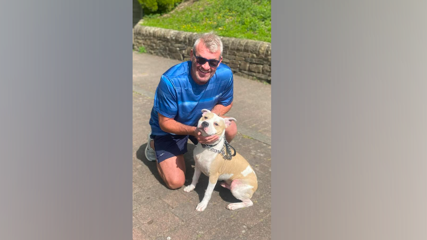 A man with grey hair, wearing black sunglasses and a blue shirt and grey shorts, kneels down on the floor, with his hands hugging a Staffordshire bull terrier type dog. It is a sunny day. 